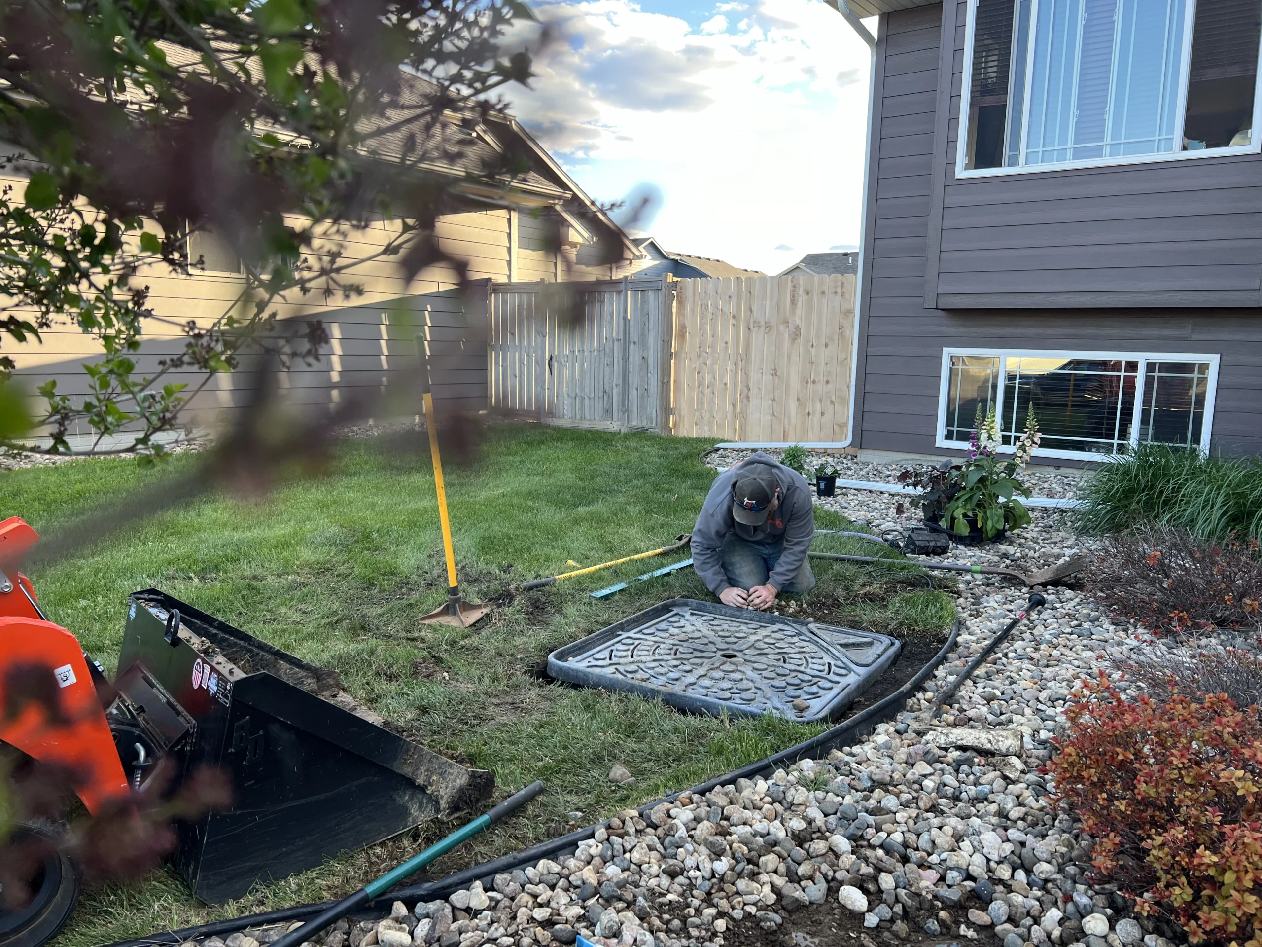 Landscaper working on a water feature installation in a residential yard, with tools and equipment visible, surrounded by grass and decorative stones.
