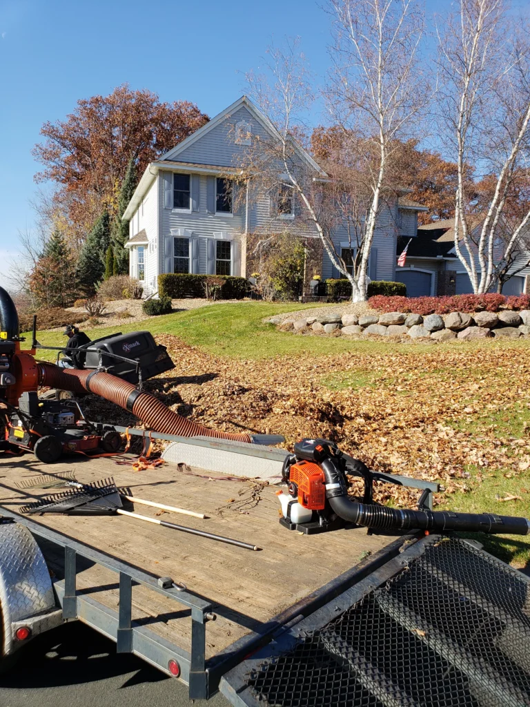 Lawn care equipment including a leaf blower on a trailer, with a suburban home in the background, showcasing autumn leaves on the ground, representing outdoor maintenance services.