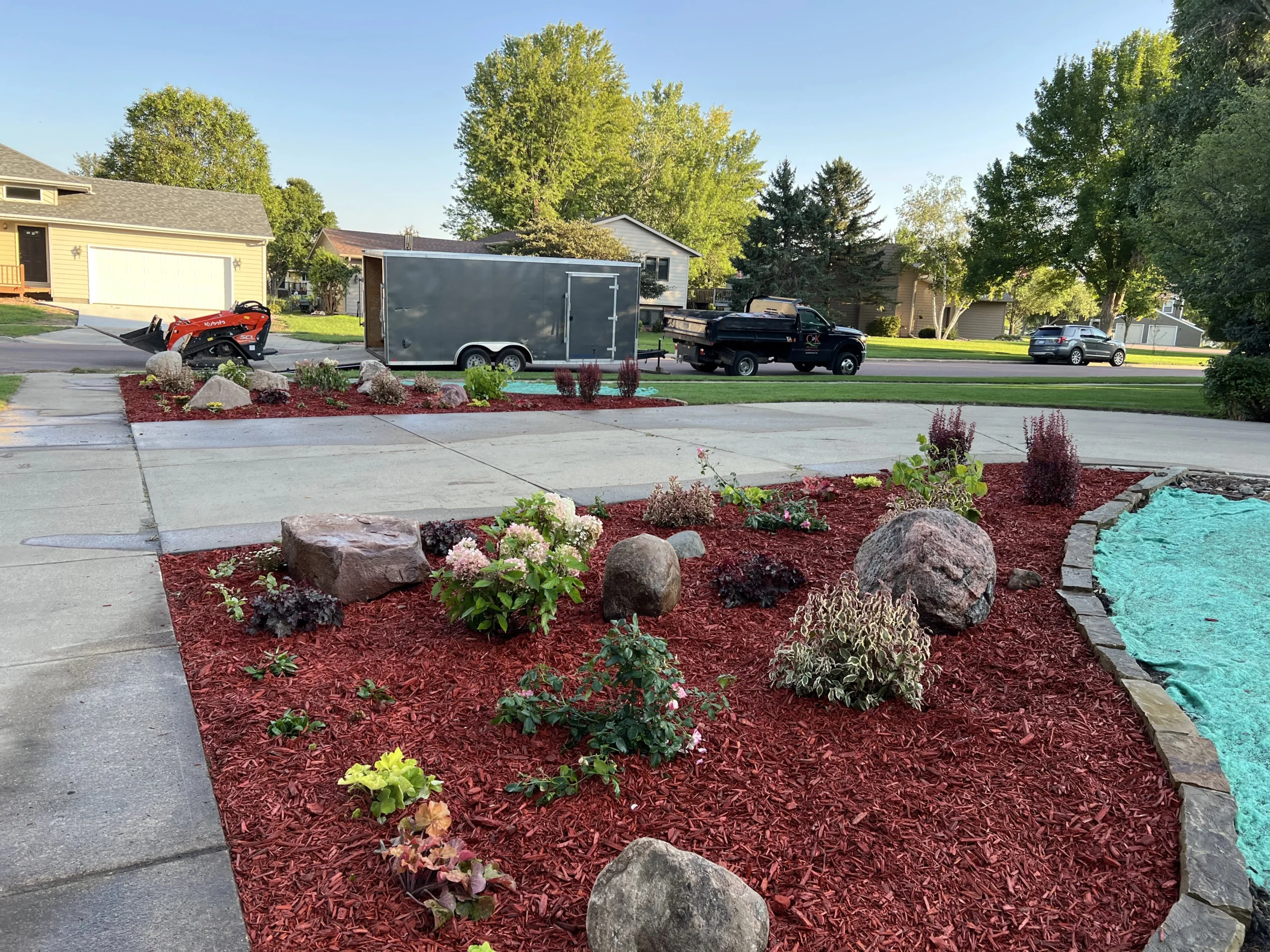 Landscaped front yard with red mulch, various plants, decorative rocks, and a landscaping trailer, showcasing professional outdoor services in Sioux Falls, SD.